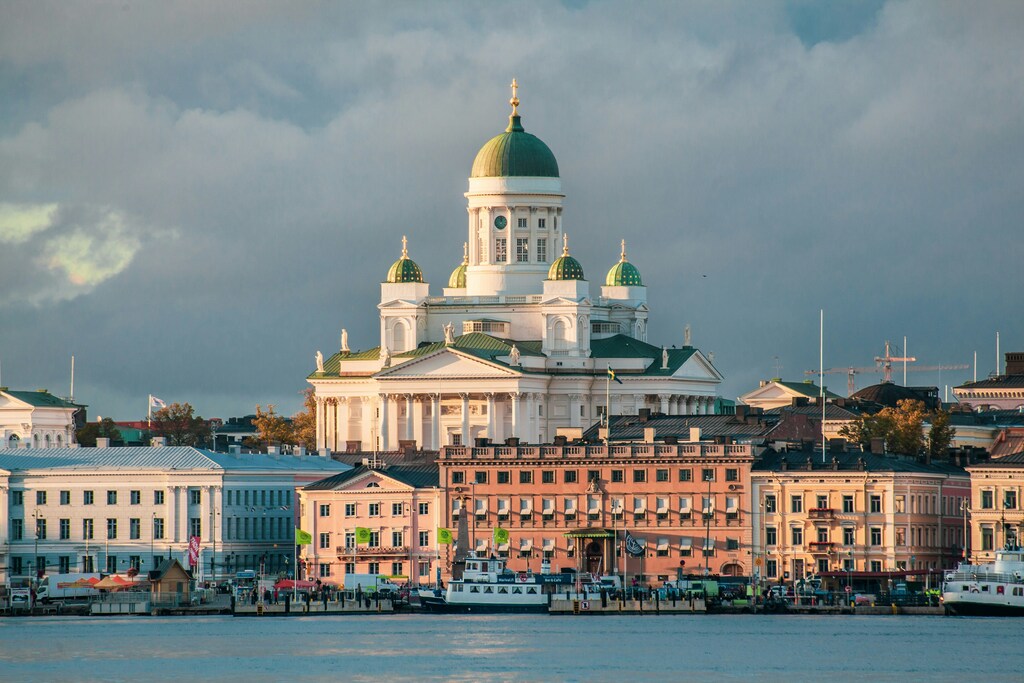 A képen a helsinki dóm impozáns fehér épülete látható a kikötő partján álló színes házak mögött.