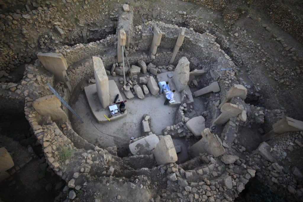 Göbeklitepe C jelű építménye feltárás és dokumentálás közben: a középen egymással szemben álló két nagy, T alakú kőoszlop két embert formáz, akiket a kőfalba ágyazottan álló, ugyancsak előre ugró fejű emberalakok vesznek körül. Írott források és részletes régészeti adatok hiányban mindennek a jelentését és funkcióját egyelőre sajnos csak találgatni lehet. (Fotó: Necmi Karul)